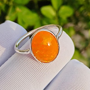 May include: A silver ring with an oval, orange gemstone. The gemstone has a vibrant, fiery colour with flecks of red and yellow. The ring is set on a white fabric background, with green foliage in the background.