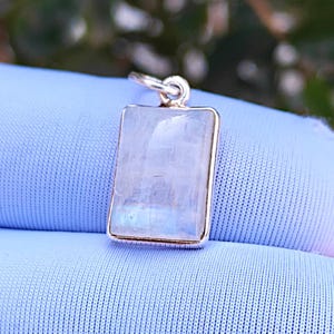 Puede incluir: Un colgante rectangular con un marco plateado y una piedra translúcida e iridiscente. La piedra tiene un sutil juego de colores, con toques de azul y blanco. El colgante está suspendido de un pequeño lazo plateado.