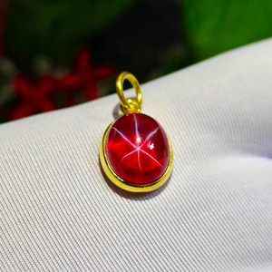 Könnte beinhalten: Ein runder, tiefroter Sternrubin-Cabochon, gefasst in einen Anhänger aus poliertem Gelbgold. Der Edelstein zeigt einen ausgeprägten sechsstrahligen Sterneffekt auf seiner glatten, gewölbten Oberfläche. Ein kleiner Goldring ist oben befestigt.
