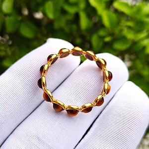 May include: A gold-colored ring with a series of small, round, amber-colored stones set around the band. The ring is displayed against a white fabric background with a blurred green foliage backdrop.