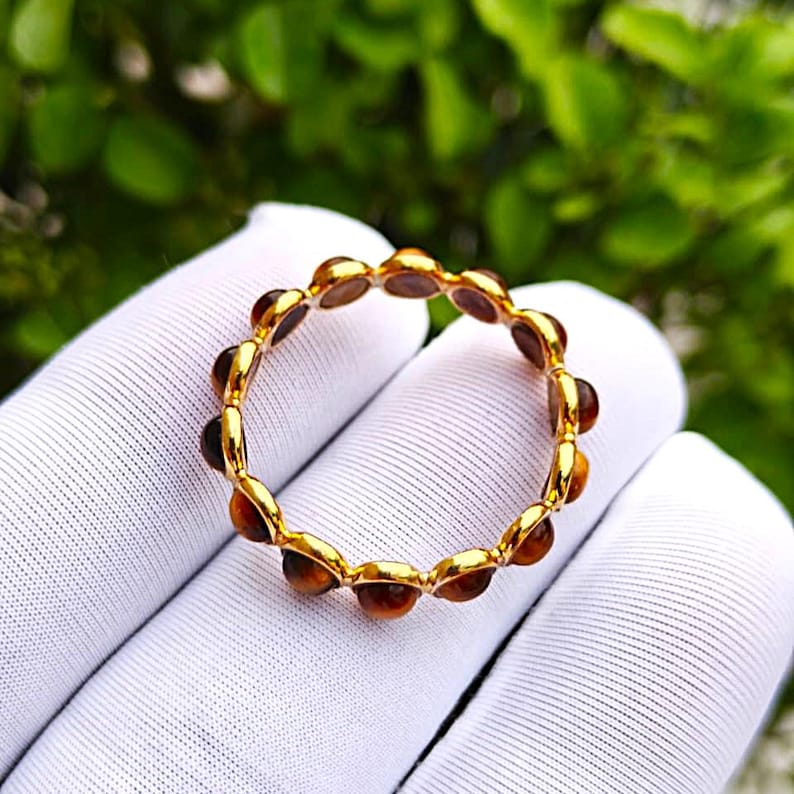 May include: A gold-colored ring with a series of small, round, amber-colored stones set around the band. The ring is displayed against a white fabric background with a blurred green foliage backdrop.