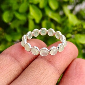May include: A silver ring featuring a series of oval-shaped, pale green gemstones set in a continuous band. The ring is displayed on a finger against a backdrop of green foliage.