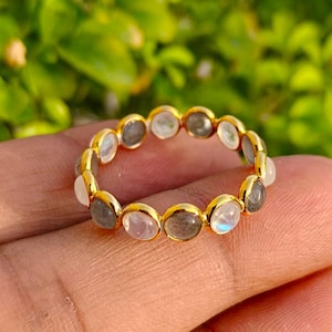 Peut inclure: Une bague en or composée d'une série de pierres rondes serties en alternance. Les pierres comprennent des gemmes translucides blanches rappelant la pierre de lune et des pierres grises opaques, formant un motif circulaire continu sur l'anneau.