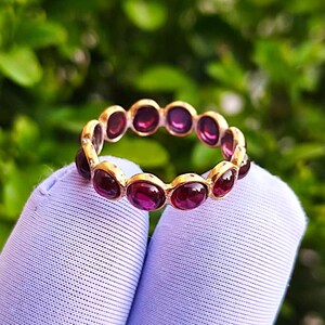 Op de afbeelding: Een gouden ring met een reeks ovale, dieprode edelstenen rond de band. De ring wordt getoond tegen een vage groene achtergrond, wat een natuurlijke omgeving suggereert. De edelstenen zijn gelijkmatig verdeeld.