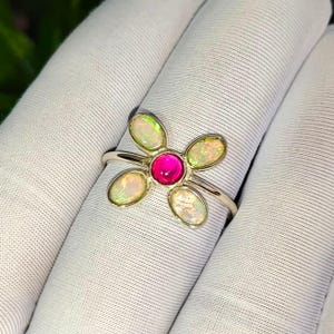 Op de afbeelding: Een zilverkleurige ring met een centrale ronde, felroze edelsteen omringd door vier ovale, iriserende opalen in een bloemvormig patroon. De metalen ringband is dun en gepolijst, wat een verfijnd bloemmotief laat zien.
