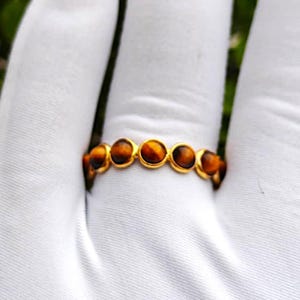 May include: A gold-colored ring featuring a series of round, tiger's eye gemstones. The gemstones are a warm brown color with golden and black streaks. The ring is set against a white background.
