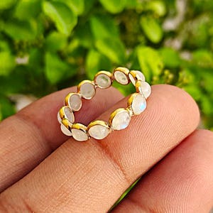 Könnte beinhalten: Ein goldfarbener Ring mit einer Reihe ovaler, schillernder Mondsteine. Der Ring ist in einem goldenen Band gefasst, und die Mondsteine sind eng um den Umfang angeordnet. Der Hintergrund ist verschwommen grün.