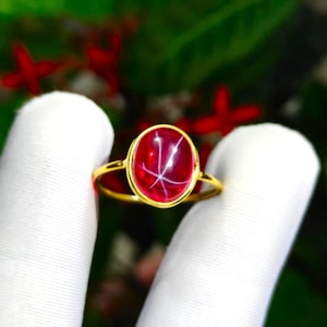 May include: A gold ring featuring a central oval cabochon-cut red gemstone. The stone displays a distinct six-rayed star pattern across its surface. The band is thin and polished, holding the gemstone in a simple bezel setting.