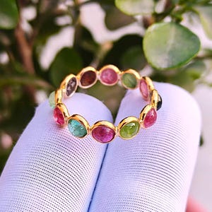 Op de afbeelding: Een gouden ring met een reeks kleurrijke edelstenen. De ring toont een verscheidenheid aan ovaal geslepen stenen in tinten rood, groen, roze en blauw. De edelstenen zijn gezet in een gouden band, waardoor een levendig en opvallend sieraad ontstaat.