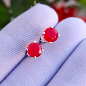 Puede incluir: Un par de pendientes de botón con piedras preciosas rojas redondas y facetadas engastadas en garras de metal plateado. Cada pendiente presenta una piedra circular de color rojo vibrante con acabado pulido, sujeta por cuatro pequeñas garras.