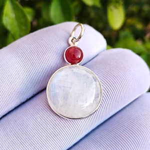May include: A round pendant with a silver bail. The pendant features a large, round, iridescent moonstone below a smaller, red gemstone. The pendant is displayed against a white background.