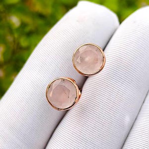 Könnte beinhalten: Ein Paar runde Ohrstecker mit facettierten, blassrosa Rosenquarz-Edelsteinen in polierten Roségold-Fassungen. Die kreisförmigen Steine haben eine glatte, mehrfach facettierte Oberfläche, die das Licht reflektiert und den Farbton betont.