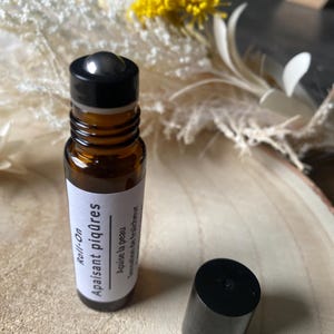 May include: A brown glass roll-on bottle with a black cap, labeled "Roll-On Apaisant piqûres" in white text. The bottle is open, with the cap beside it. The background includes dried flowers and a wooden surface.