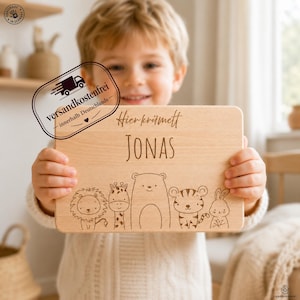 May include: A rectangular light-colored wooden cutting board featuring the engraved text "Hier krümelt" above the name "JONAS" and a series of five illustrated animal characters including a lion, giraffe, bear, tiger, and rabbit.