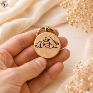 Könnte beinhalten: Ein runder Schlüsselanhänger aus hellem Holz mit einem silbernen Schlüsselring. Der Anhänger zeigt eine schwarze Illustration von zwei Fäusten, die sich berühren, mit den Worten "LEON" und "PAPA" auf beiden Seiten.