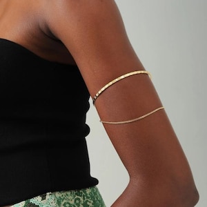 May include: Gold arm cuff jewellery. The upper band has a textured design, while the lower band is a delicate chain. The jewellery is worn on the upper arm. The person is wearing a black top and green patterned trousers.