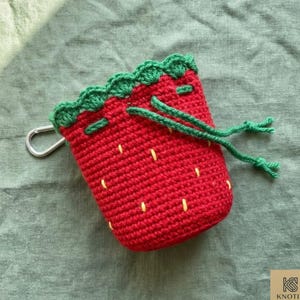 May include: A handmade red crochet pouch shaped like a strawberry, with green trim and a drawstring closure. The pouch has yellow seed details and a silver carabiner clip. The pouch is on a green surface.