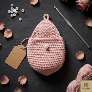Könnte beinhalten: Eine handgefertigte, hellrosa gehäkelte Tasche mit Perlenakzent. Auf einem dunkelgrauen Hintergrund liegen Rosenblätter, Knöpfe und eine Häkelnadel. Ein leeres Etikett deutet darauf hin, dass es sich um ein Geschenk oder einen Artikel zum Verkauf handelt.