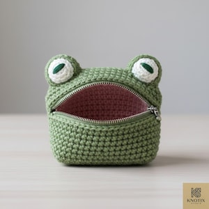 May include: A green crocheted frog-shaped pouch with a zipper closure. The frog has white and green eyes and a pink interior. The pouch is designed to resemble a frog's open mouth.