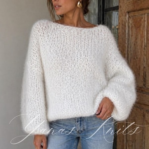 Puede incluir: Un suéter blanco de punto de gran tamaño con cuello barco y mangas largas. El suéter tiene una apariencia esponjosa y texturizada. El texto "Yanas Knits" es visible en la esquina inferior izquierda.