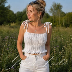 May include: A white crochet crop top featuring a ribbed texture, a ruffled hem, and adjustable shoulder straps tied in bows. The garment is paired with white denim jeans. The text Yanas Knits is visible in a cursive script at the bottom.