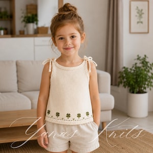 May include: A cream-coloured sleeveless knit top featuring tie-shoulder straps and a decorative green embroidered clover pattern along the bottom hem. The garment is paired with light beige shorts, creating a coordinated and simple outfit.
