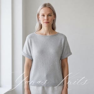 Puede incluir: Una blusa de punto gris claro de manga corta con un amplio escote barco, mangas con dobladillo y un dobladillo acanalado. La prenda tiene una textura fina y suave con un corte recto y relajado. El texto "Yanas Knits" está superpuesto en letra cursiva blanca.
