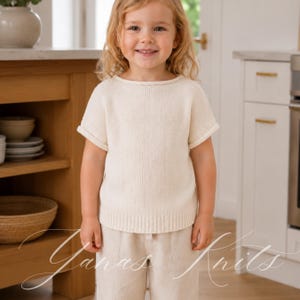 May include: A short-sleeved, cream-coloured knit jumper featuring a round neckline and ribbed trim at the hem and sleeve cuffs. The garment is constructed with a simple stockinette stitch pattern, providing a clean and uniform texture throughout.
