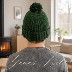 May include: A dark forest green knit beanie featuring a ribbed cuff and a matching pom-pom on top. The hat is worn on the head, and the text Yanas Knits is written in a white cursive script across the lower portion of the image.
