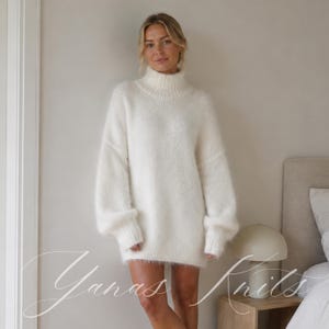 May include: An oversized, long-sleeved, white knit jumper featuring a high turtleneck collar and a soft, fuzzy texture. The garment has dropped shoulders and ribbed cuffs at the wrists and hem, providing a relaxed fit that reaches mid-thigh.