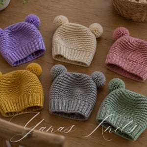 May include: Six knitted baby hats in a range of colours: purple, yellow, grey, pink, cream, and green. Each hat has a pom-pom on top and two smaller pom-poms on the sides, resembling bear ears. The hats are displayed on a wooden surface.