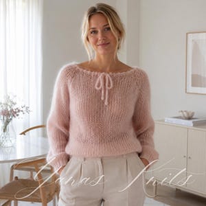 May include: A pale pink long-sleeved sweater crafted from a fuzzy, mohair-style knit fabric. The garment features a wide, rounded neckline with a delicate, matching knitted tie detail at the center and long sleeves with ribbed cuffs.