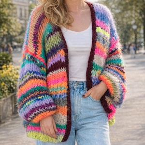 Può includere: Un cardigan oversize e vibrante in un arcobaleno di colori. Il maglione aperto presenta strisce viola, arancioni, rosa, blu e verdi. Le maniche sono lunghe e voluminose e il bordo è bordeaux scuro.