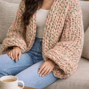 Könnte beinhalten: Ein grob gestrickter Cardigan in einer weichen Farbpalette aus Rosa, Beige und Grün. Der übergroße Pullover hat weite Ärmel und eine lockere Passform. Das Model trägt den Cardigan mit einem weißen Oberteil und blauen Jeans. Eine weiße Kaffeetasse steht auf einem Tisch.