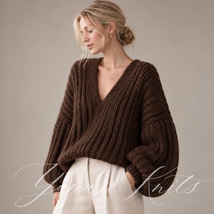 May include: A dark brown, chunky knit jumper featuring a deep V-neckline and voluminous long sleeves with ribbed cuffs. The jumper is paired with light beige trousers. The text "Yanas Knits" is written in a white cursive script across the image.