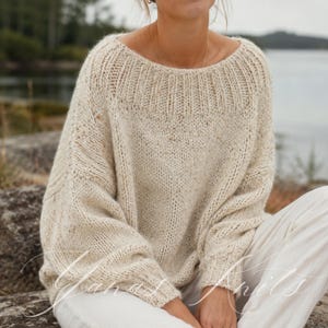 Puede incluir: Un suéter de punto crema de gran tamaño con un escote amplio. El suéter tiene una apariencia texturizada con motas de hilo más oscuro. Las mangas son largas y el estilo general es relajado y cómodo. Se ve un pantalón blanco.