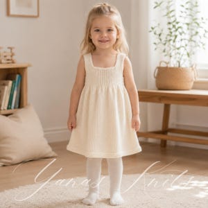 May include: A sleeveless, cream-colored knit dress featuring a square neckline and a flared skirt. The garment is crafted with a visible stockinette stitch pattern throughout the bodice and skirt, finished with a simple ribbed hem at the bottom edge.