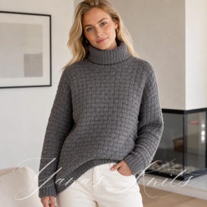 May include: A charcoal gray long-sleeve sweater featuring a prominent basketweave knit texture, a high turtleneck collar, and ribbed cuffs. The garment is paired with white pants. The text Yanas Knits is visible in white cursive script across the lower portion.