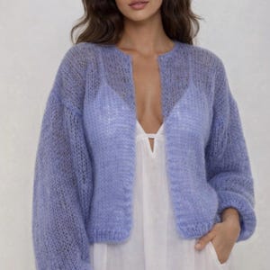 Puede incluir: Un cárdigan azul lavanda claro con mangas largas. El suéter abierto está hecho de un material de punto suave y transparente. El cárdigan tiene un corte holgado y un cuello redondo.