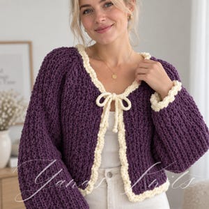 May include: A deep purple chunky knit cardigan featuring long sleeves and a contrasting cream-colored trim along the neckline, front opening, and cuffs. The garment includes a matching cream-colored tie closure at the center of the chest.
