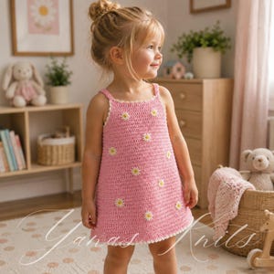 Puede incluir: Un vestido de ganchillo rosa sin mangas con un patrón repetitivo de pequeñas margaritas blancas con centros amarillos. El vestido tiene una silueta sencilla en forma de A con un borde festoneado blanco a lo largo del cuello y el dobladillo.