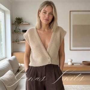Può includere: Un gilet smanicato in maglia beige con profondo scollo a V e chiusura frontale con due bottoni visibili. Il capo presenta una consistenza morbida e soffice e una lunghezza corta, abbinato a pantaloni marrone scuro. È visibile il testo Yanas Knits.