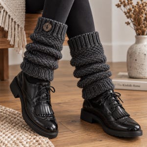 Peut inclure: Une paire de jambières en tricot gris anthracite foncé avec une texture côtelée et un bouton rond en bois sur le revers supérieur, portées sur des leggings noirs et associées à des chaussures richelieu à lacets en cuir noir à franges.
