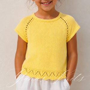 May include: A bright yellow knitted top with short sleeves. The top features a decorative openwork design along the neckline and hem. The garment is made of a soft, textured yarn, perfect for a child's summer wardrobe.