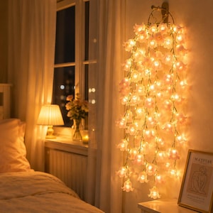 Puede incluir: Una escena de dormitorio con una cama, una ventana y luces de cadena decorativas. Las luces de cadena son pequeñas luces en forma de flor rosa que cuelgan de un accesorio de pared. Una impresión enmarcada y un jarrón de flores están en una mesita de noche.