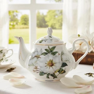 Puede incluir: Tetera de porcelana blanca con un diseño floral de magnolia. La tetera presenta un pico curvo, un asa de bucle y una tapa con un adorno en forma de capullo. Los bordes están adornados con ribetes dorados. La tetera está sobre un mantel blanco.