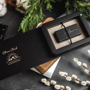 Può includere: Un'unità USB nera in una scatola nera con interno in lino, appoggiata su una superficie in legno. La scatola fa parte di un folio nero con il nome "Olivia Clark Photographer" e un logo di montagna. Elementi decorativi: una fotocamera vintage e ramoscelli.