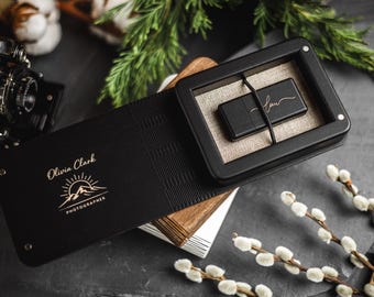May include: A black USB drive in a black box with a linen interior, resting on a wooden surface. The box is part of a black folio with the name "Olivia Clark Photographer" and a mountain logo. Decorative elements include a vintage camera and sprigs.
