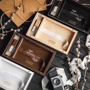 Puede incluir: Vista aérea de cajas de presentación de madera, algunas abiertas, con una cámara vintage y elementos decorativos. Las cajas son de color negro, marrón y madera natural. Algunas cajas tienen cintas y el texto "WEDDING DAY".