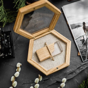 May include: A hexagonal wooden box with a glass lid featuring the engraved names Daniel and Emma. Inside, a rectangular wooden and clear resin USB flash drive rests on a beige fabric lining, secured by a thin wooden bar across the center.